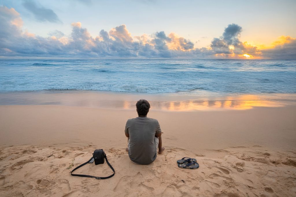 pexels-photo-30481618-30481618 Man sitting on sandy beach at sunset, gazing at the ocean, reflecting a peaceful sunset.